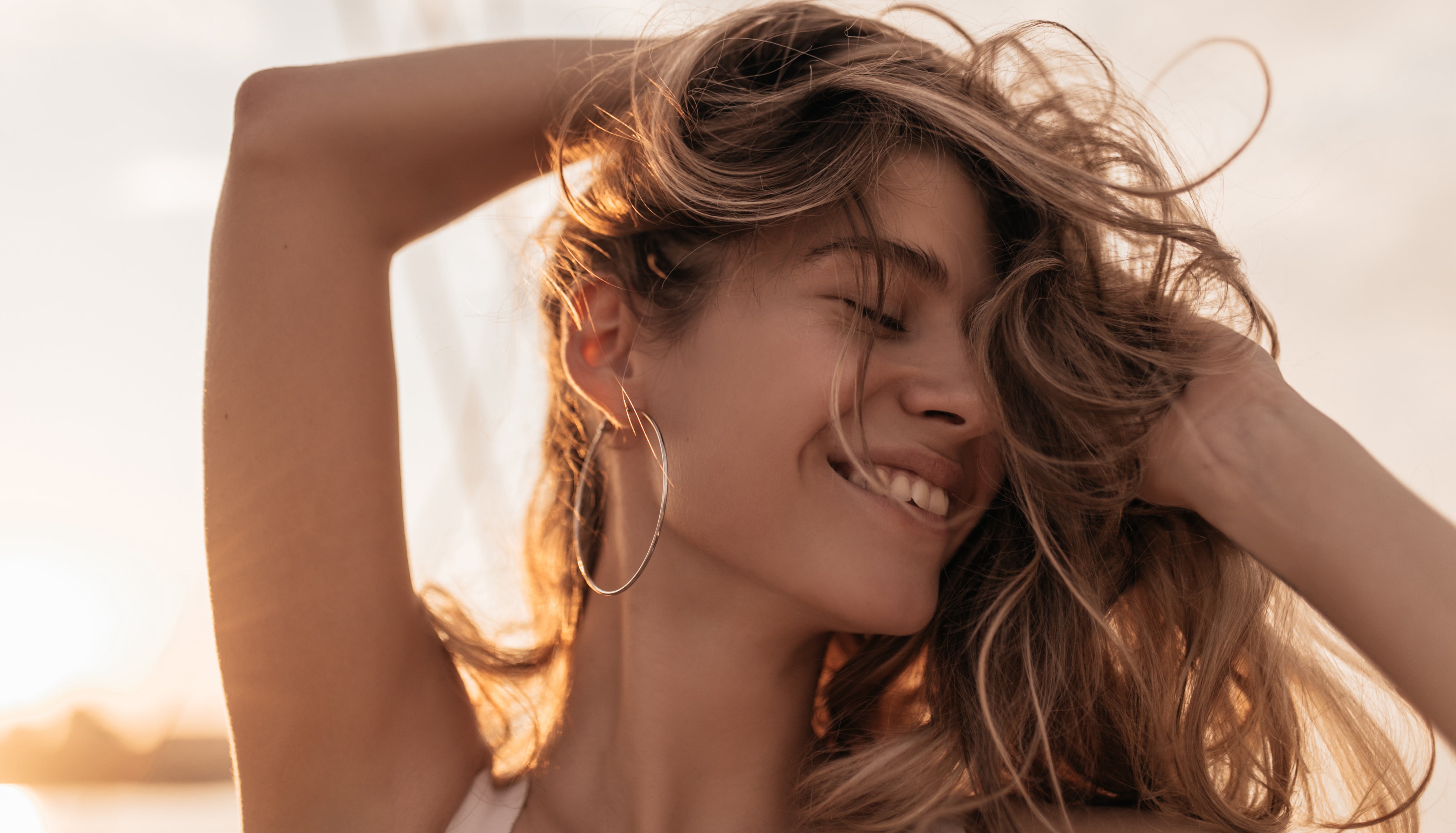A smiling woman with long, wavy, sun-kissed blonde hair running her fingers through her healthy, flowing locks, symbolizing hair beauty and care.