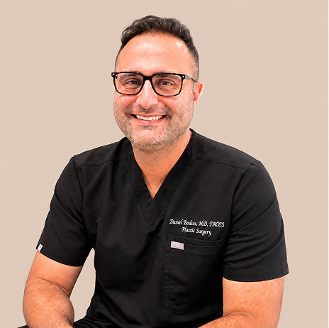 Dr. David Kashan, MD, FACS, a plastic and reconstructive surgeon, smiling in black scrubs.