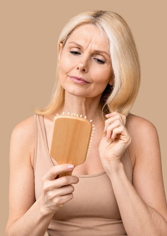 A concerned woman with blonde hair looking at strands of shed hair caught in her wooden paddle brush.